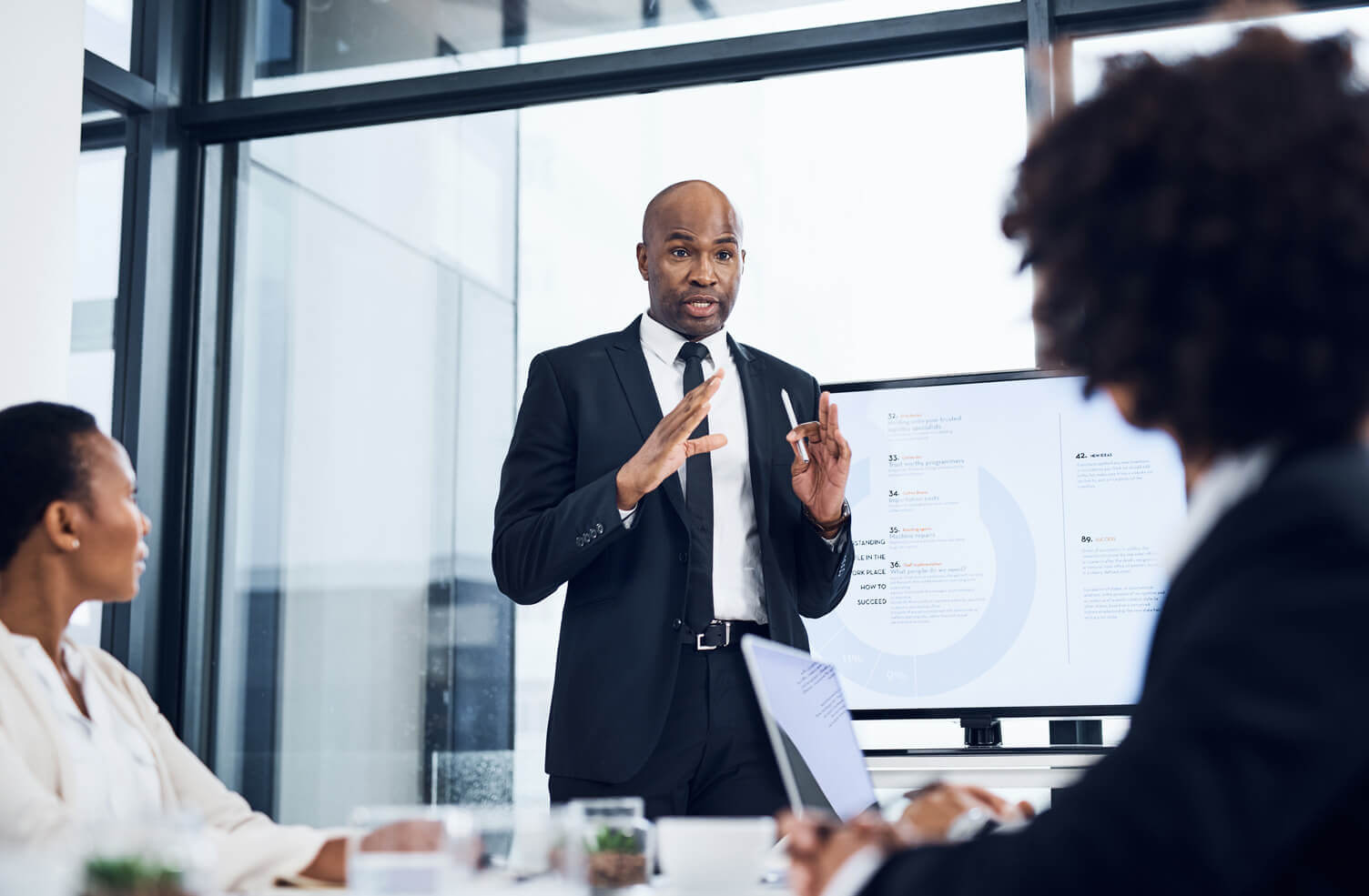 a man talking in a meeting, behind him is a large screen with a pi-chart on it